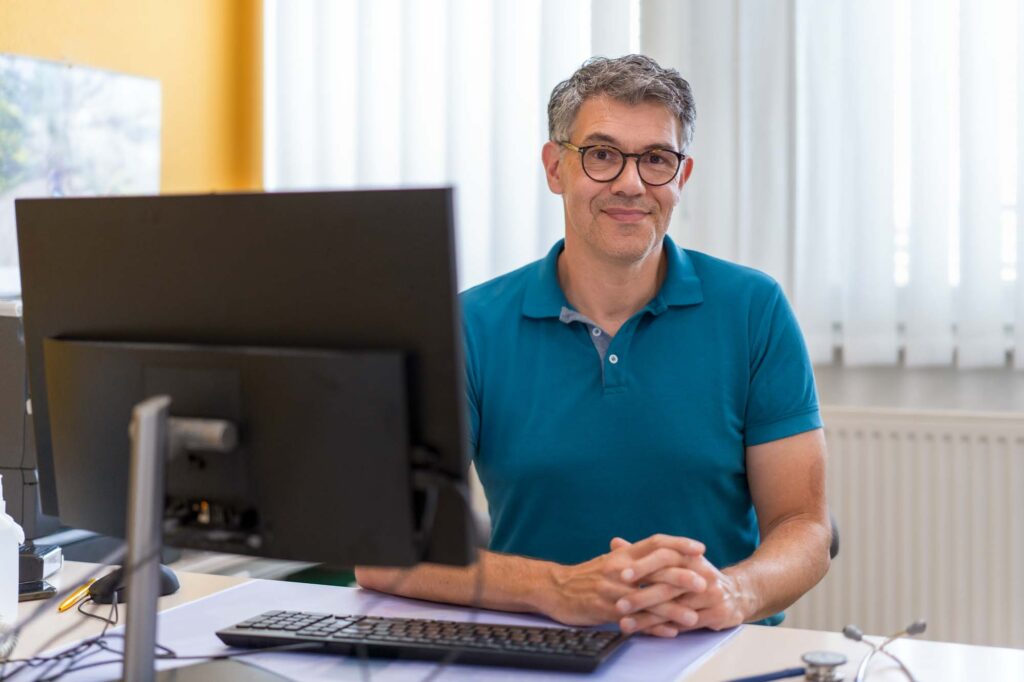 Dr. Dimos sitzt im Sprechzimmer am Schreibtisch und blickt freundlich in die Kamera